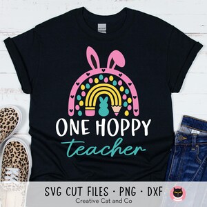 Pu&ograve; includere: Maglietta nera con un arcobaleno, orecchie di coniglio e un design a matita. Il testo "One Hoppy Teacher" &egrave; stampato sulla maglietta.
