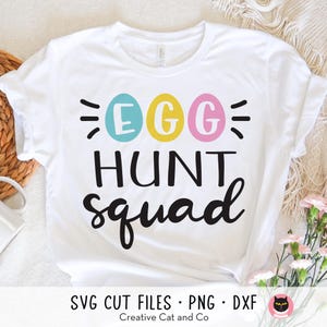 Puede incluir: Camiseta blanca con el texto "Egg Hunt Squad" en negro. Tres huevos de colores están por encima del texto. Los huevos son azul, amarillo y rosa.