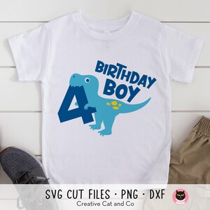 May include: A white t-shirt with a blue dinosaur holding a blue number four. The text "Birthday Boy" is above the dinosaur.