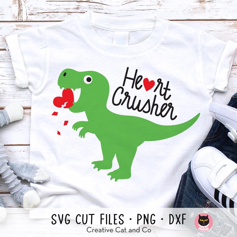 May include: White t-shirt with a green dinosaur graphic and the text "Heart Crusher" in black. The dinosaur is holding a red heart in its mouth.