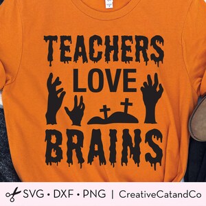 K&ouml;nnte beinhalten: Orangefarbenes T-Shirt mit schwarzem Text, der "Teachers Love Brains" mit einem Tropfeffekt lautet. Der Text ist von schwarzen Zombie-Handsilhouetten und einem kleinen Friedhof mit zwei Kreuzen umgeben.