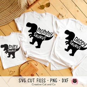 May include: Three white t-shirts with black dinosaur silhouettes and text. The shirts read "Mommy Saurus", "Daddy Saurus", and "Mini Saurus".