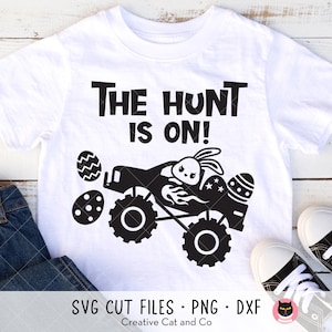 May include: White t-shirt with the text "THE HUNT IS ON!" in black. A black graphic depicts a monster truck with Easter eggs and a bunny. The shirt is paired with blue jeans and white sneakers.