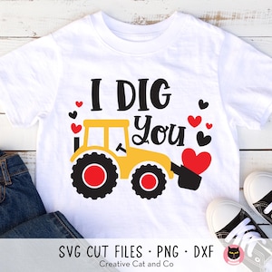 May include: A white t-shirt with a yellow tractor design and the text "I DIG you" in black. There are red hearts scattered around the tractor. The shirt is on a white wooden background.
