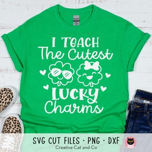 May include: Green t-shirt with white text that reads "I Teach The Cutest Lucky Charms". The text is surrounded by two shamrock designs, one with sunglasses and the other with a bow.