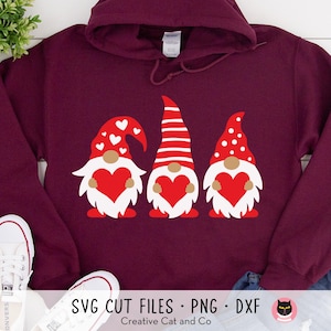 May include: A maroon hoodie with a design of three gnomes holding hearts. The gnomes are wearing red and white hats. The text "SVG CUT FILES: PNG - DXF" is at the bottom of the image. The text "Creative Cat and Co" is below the text.