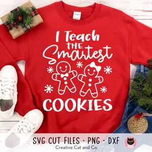 May include: Red sweatshirt with white text that reads "I Teach the Smartest Cookies". The text is surrounded by white snowflakes and two gingerbread people.