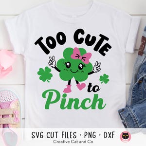 May include: A white t-shirt with a green shamrock design and the text "Too Cute to Pinch". The shamrock has a pink bow and a smiling face.