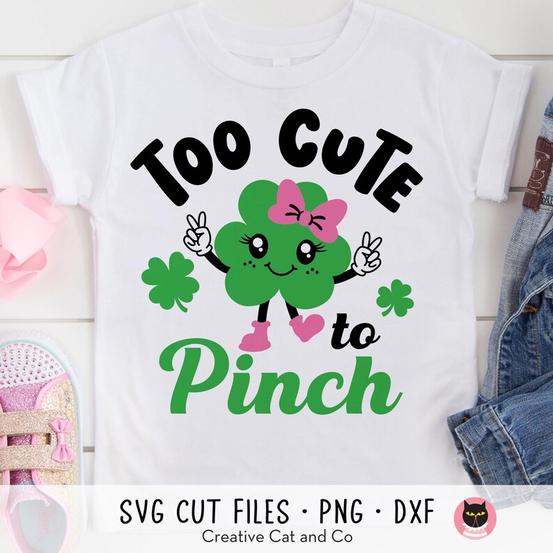May include: A white t-shirt with a green shamrock design and the text "Too Cute to Pinch". The shamrock has a pink bow and a smiling face.