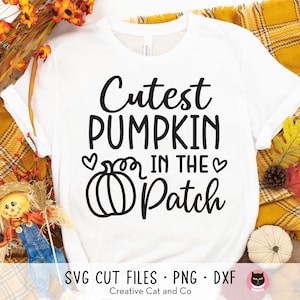 May include: White t-shirt with black text that reads "Cutest Pumpkin in the Patch" and a black outline of a pumpkin. The shirt is laying on a plaid fabric background.