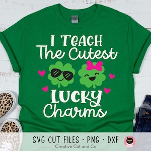 Puede incluir: Camiseta verde con el texto "I Teach The Cutest Lucky Charms" en blanco. Dos tréboles de dibujos animados con caras sonrientes están en la camiseta.