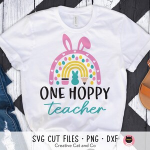 Pu&ograve; includere: Maglietta bianca con un design a forma di arcobaleno con uova di Pasqua, matite e un coniglio. Il testo "ONE HOPPY teacher" &egrave; stampato sotto l'arcobaleno.