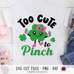 May include: A white t-shirt with a green shamrock design and the text "Too Cute to Pinch". The shamrock has a pink bow and a smiling face.