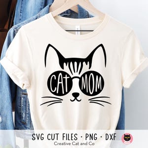 Pode incluir: Uma camiseta branca com um gato preto usando óculos de sol e o texto "Cat Mom".