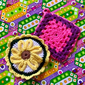 May include: Two handmade crochet items: a pink and purple square potholder and a yellow, white, and green sunflower-shaped coaster. The items are on a colorful patterned fabric background with green, yellow, purple, and white geometric shapes.