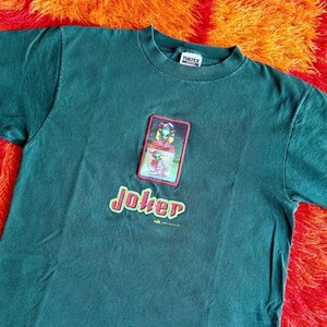 May include: Dark green t-shirt featuring a graphic of a character and the word "Joker" in a red and green font. The shirt has a crew neck and a small tag at the neckline.