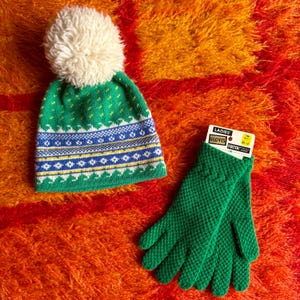May include: A green knit hat with a white pom-pom and a matching pair of green gloves. The hat has a blue and yellow patterned design. The gloves have a textured pattern and a tag that reads "LADIES GLOVES STRETCH".