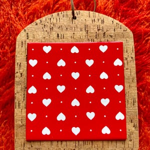 May include: A decorative corkboard with a red background featuring a pattern of white hearts. The corkboard has a natural wood grain frame and a brown leather loop for hanging. The overall design is simple and charming.