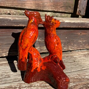 May include: Ceramic sculpture of two birds perched on a tree stump. The birds are painted in shades of orange and red with a glossy finish.