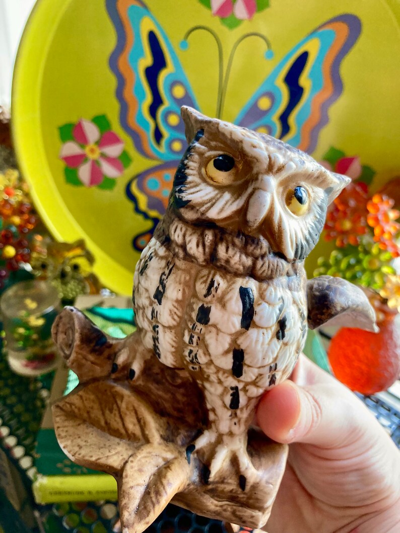 Op de afbeelding: Een keramiek uilenbeeldje dat op een tak zit, met bruine en witte veren en grote gele ogen. De uil kijkt naar links en de tak is bruin en beige geverfd.