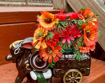 Op de afbeelding: Een bruine keramische ezel die een wagen-plantenbak trekt met een boeket oranje en rode bloemen. De wagen heeft de vorm van een bruin vat met een gouden wiel.