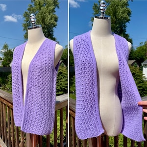 May include: A sleeveless lavender knit vest featuring a textured waffle-like pattern throughout the fabric. The garment has an open front design with ribbed trim along the edges and armholes, providing a consistent look across the entire piece.