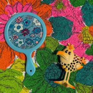 May include: A light blue hand mirror with a floral design and a vintage bird brooch. The bird brooch is cream-colored with orange and yellow accents. The background is a colorful floral pattern.