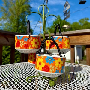May include: A set of four hanging ceramic planters with a vibrant floral design. The planters feature an orange background with yellow, blue, and red flowers. Each planter has a black handle and is suspended from a green metal stand.