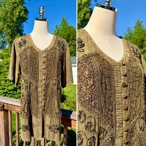 May include: A short-sleeved olive green tunic featuring a V-neckline and a front button placket. The garment is decorated with intricate dark brown embroidery, textured fabric panels, and swirling patterns across the front and shoulder areas.