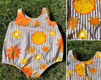 May include: A white and brown striped baby onesie with a yellow and orange daisy print. The onesie has a sleeveless design and says "Daisy" on the front.