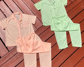May include: Two sets of pajamas in the image. One set is a light peach color, and the other is a light green. Each set includes a short-sleeved button-down shirt with a chest pocket and matching pants. The pajamas are made of a soft, lightweight fabric.