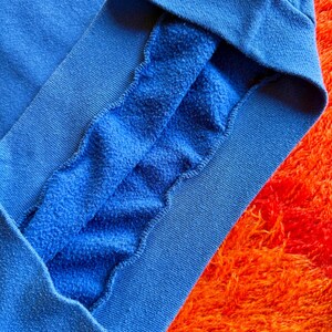 Pu&ograve; includere: Primo piano di una manica di felpa blu, che rivela l'interno morbido e felpato. Il tessuto blu vibrante contrasta con lo sfondo arancione strutturato. Il polsino &egrave; di una tonalit&agrave; di blu pi&ugrave; scura.