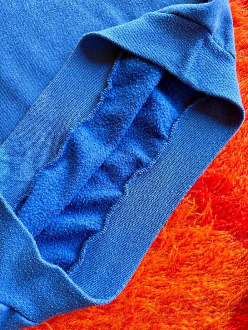Pu&ograve; includere: Primo piano di una manica di felpa blu, che rivela l'interno morbido e felpato. Il tessuto blu vibrante contrasta con lo sfondo arancione strutturato. Il polsino &egrave; di una tonalit&agrave; di blu pi&ugrave; scura.
