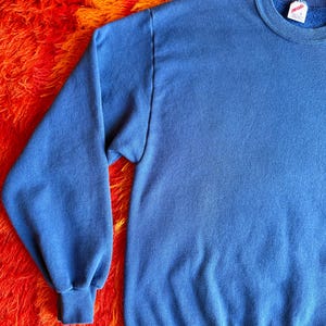 Pu&ograve; includere: Primo piano di una felpa blu. La parte superiore a maniche lunghe ha un girocollo e polsini e orlo a coste. Il tessuto appare morbido e leggermente usurato. Lo sfondo &egrave; un vivace tappeto shag arancione e rosso.