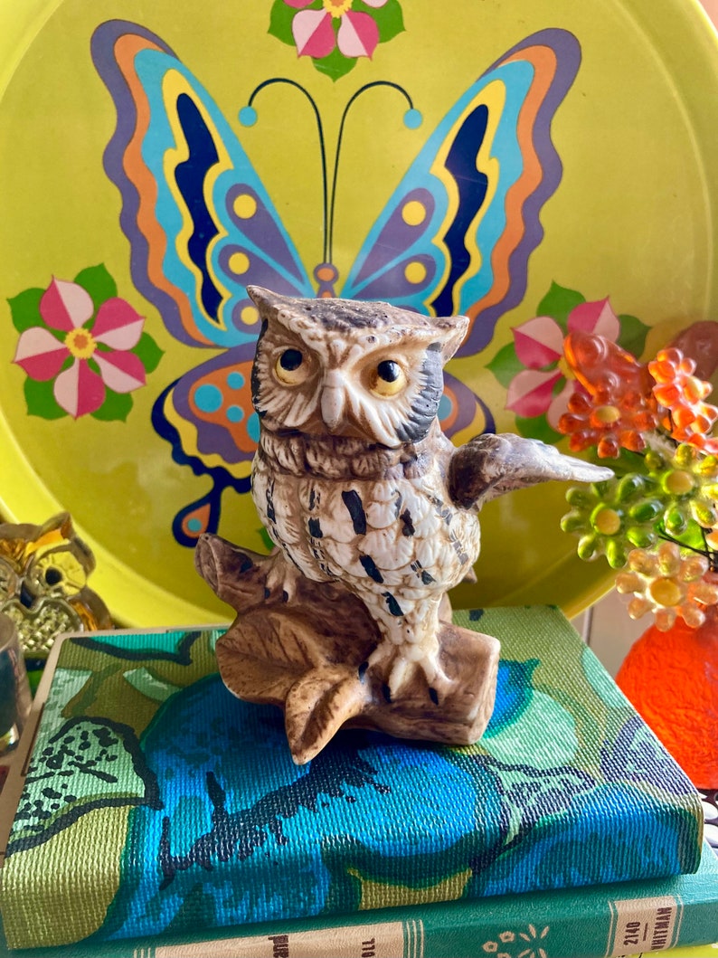 Op de afbeelding: Een keramiek uilbeeldje met bruine en witte markeringen, gepercht op een tak met groene bladeren. De uil heeft gele ogen en kijkt naar rechts. Het beeldje staat op een boek met een blauw en groen bloemenpatroon.