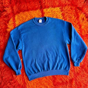 Pu&ograve; includere: Una felpa blu a girocollo tinta unita. La parte superiore a maniche lunghe ha polsini e orlo a coste. La felpa &egrave; realizzata in un materiale morbido ed &egrave; esposta su un vivace tappeto shaggy arancione e giallo.