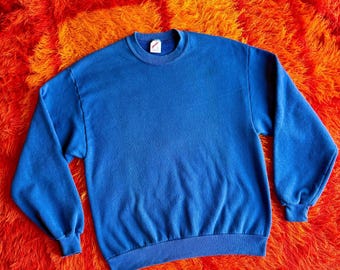 May include: A solid blue crew neck sweatshirt. The long-sleeved top has ribbed cuffs and a ribbed hem. The sweatshirt is made of a soft material and is displayed on a vibrant orange and yellow shag carpet.