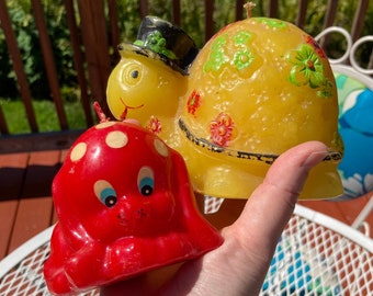 Op de afbeelding: Twee vintage kaarsfiguurtjes. Een gele schildpad met een zwarte hoed en groene bloemen en een rode puppy met witte stippen.
