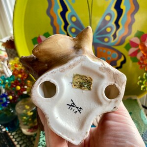 Op de afbeelding: Een wit keramiek eekhoornbeeldje met bruine beschilderde details. Het beeldje staat op zijn achterpoten met zijn staart opgerold. De onderkant van het beeldje heeft een label met de tekst "HOMCO MADE IN TAIWAN" en een nummer "1114".