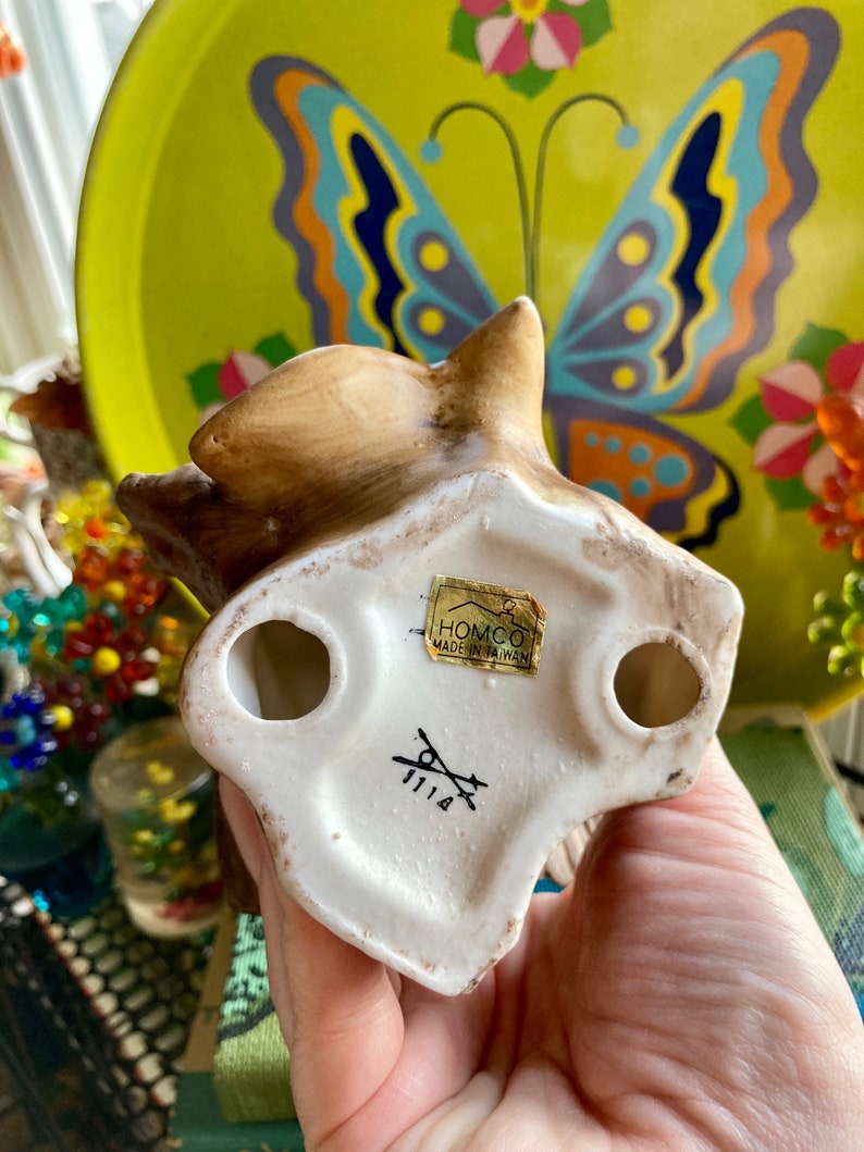 Op de afbeelding: Een wit keramiek eekhoornbeeldje met bruine beschilderde details. Het beeldje staat op zijn achterpoten met zijn staart opgerold. De onderkant van het beeldje heeft een label met de tekst "HOMCO MADE IN TAIWAN" en een nummer "1114".