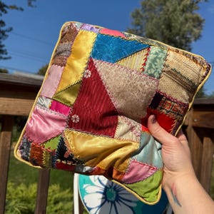 May include: A colorful patchwork pillow with a variety of fabrics in shades of red, yellow, blue, and green. The pillow has a yellow border and is held up against a blue sky.