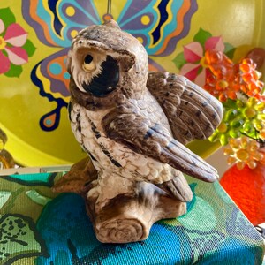 Op de afbeelding: Een keramiek uilenbeeldje met bruine en witte markeringen, geperst op een boomstronk. De uil kijkt naar links, met zijn vleugels uitgespreid. Het beeldje staat op een stapel boeken met kleurrijke omslagen.