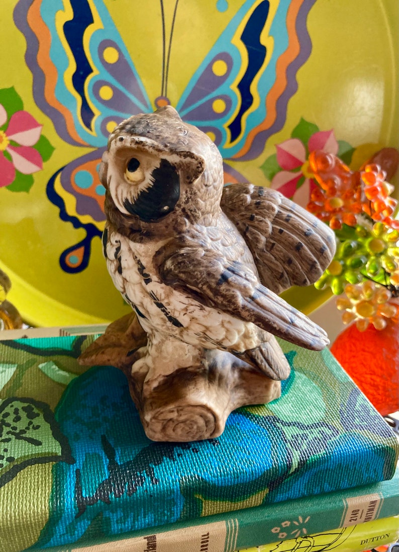 Op de afbeelding: Een keramiek uilenbeeldje met bruine en witte markeringen, geperst op een boomstronk. De uil kijkt naar links, met zijn vleugels uitgespreid. Het beeldje staat op een stapel boeken met kleurrijke omslagen.