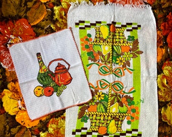 May include: Two white kitchen towels with a fringe border. The towels feature a colorful fruit and basket design. One towel has a teapot, fruit, and grapes. The other towel has a basket of fruit with a green and brown checkered border.