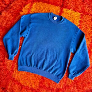 Pu&ograve; includere: Una felpa blu tinta unita con girocollo e maniche lunghe. La felpa &egrave; realizzata in un materiale morbido e viene esposta su un vivace tappeto shag arancione e rosso. L'etichetta sul colletto &egrave; visibile.
