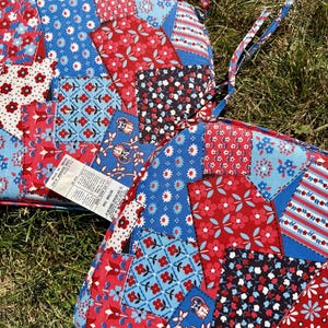 May include: Two square seat cushions featuring a patchwork design with various floral and geometric patterns in red, white, and blue. Each cushion has fabric ties at the corners and a white product tag attached with text detailing material content.