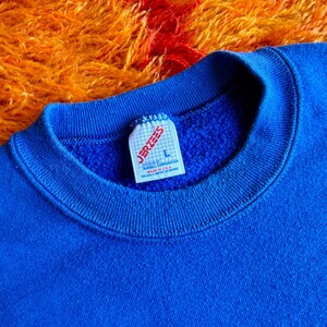 Pu&ograve; includere: Primo piano di una felpa blu reale con scollo tondo. L'etichetta recita "JERZEES" e "L". Lo sfondo &egrave; una superficie strutturata arancione e rossa. La felpa &egrave; realizzata in un materiale morbido.