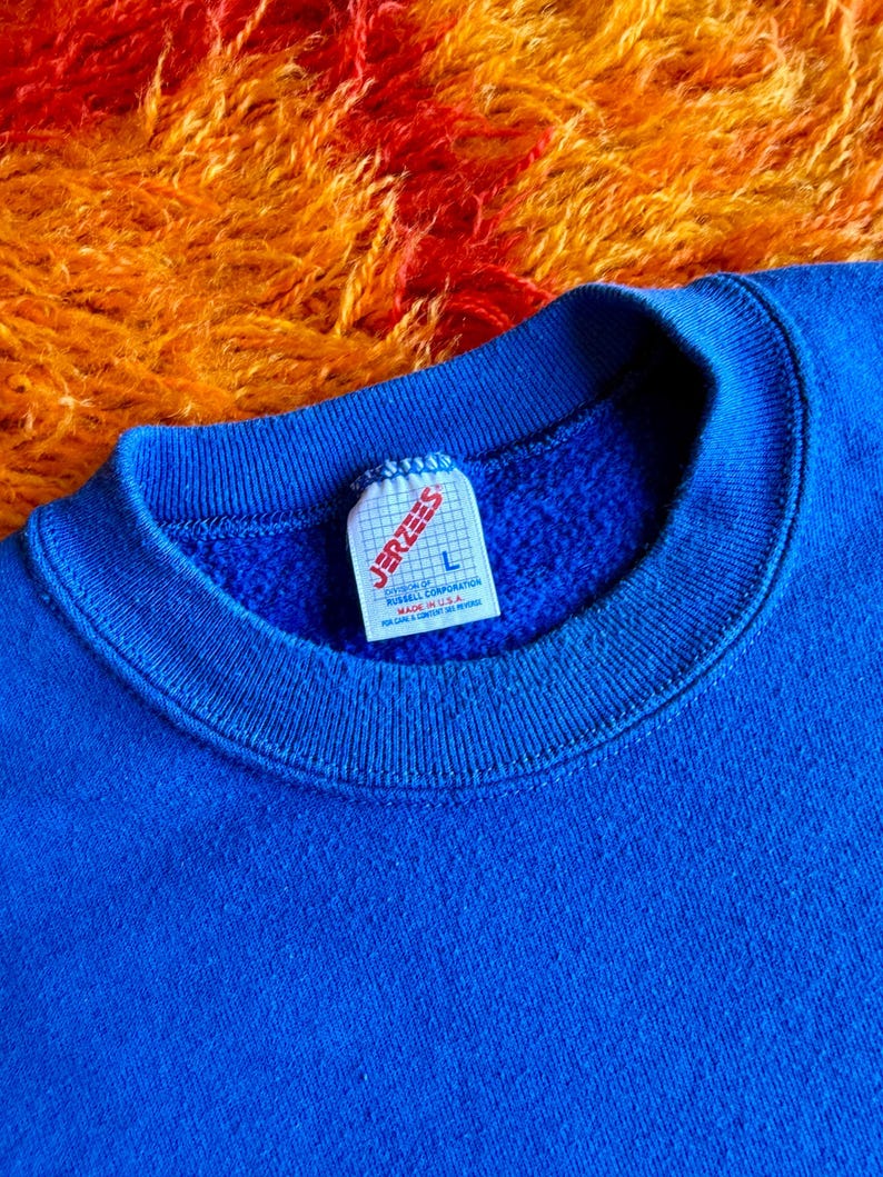 Pu&ograve; includere: Primo piano di una felpa blu reale con scollo tondo. L'etichetta recita "JERZEES" e "L". Lo sfondo &egrave; una superficie strutturata arancione e rossa. La felpa &egrave; realizzata in un materiale morbido.