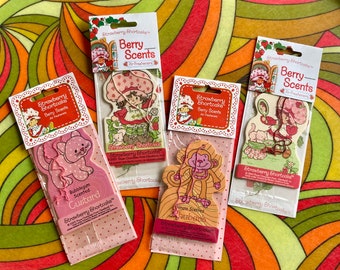 May include: A set of ten Strawberry Shortcake air fresheners. Each air freshener features a different Strawberry Shortcake character and a different scent. The scents include bubblegum, custard, banana, and strawberry. The air fresheners are packaged in colorful envelopes with the Strawberry Shortcake logo.