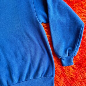 Pu&ograve; includere: Primo piano di una felpa blu reale. La felpa ha maniche lunghe con polsini e orlo a coste. Il tessuto sembra essere un materiale morbido e confortevole. Lo sfondo &egrave; una superficie arancione brillante e strutturata.